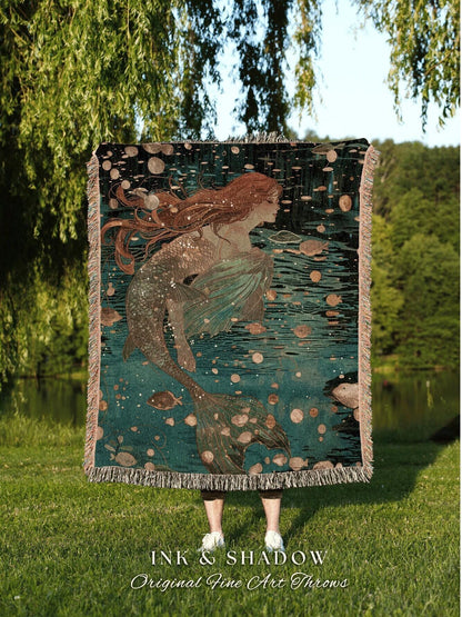 May include: A woven throw blanket featuring a mermaid illustration in shades of teal, brown, and beige. The mermaid has long red hair and a flowing tail, swimming in a dark teal water with bubbles and fish. The blanket has a fringed edge. Text at the bottom reads "INK & SHADOW Original Fine Art Throws."