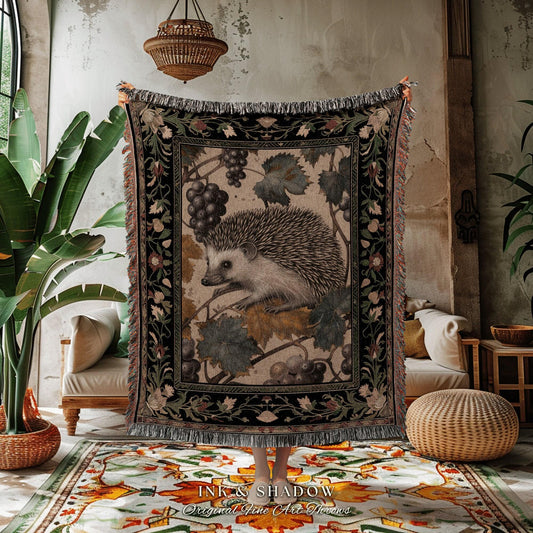 May include: A tapestry throw blanket featuring a detailed illustration of a hedgehog amidst grape vines. The throw has a black border with floral accents and fringed edges. The text "INK & SHADOW Original Fine Art Throws" is at the bottom.