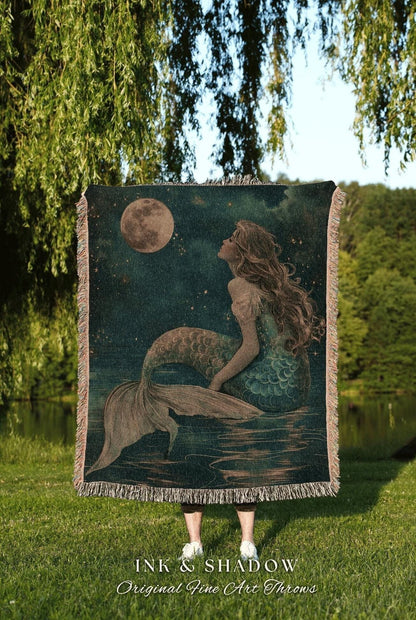 May include: A woven throw blanket featuring a mermaid illustration under a full moon in a starry night sky. The mermaid has long flowing hair and a teal tail. The blanket has a fringed edge and the text "INK & SHADOW Original Fine Art Throws".