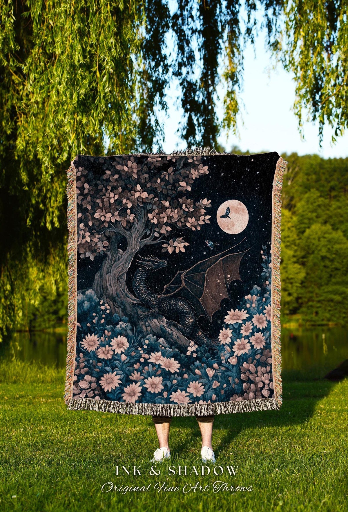 May include: A woven throw blanket featuring a fantasy scene with a black dragon, a full moon, and a tree with pink and white flowers. The blanket has a fringed edge and the text "INK & SHADOW Original Fine Art Throws" at the bottom.