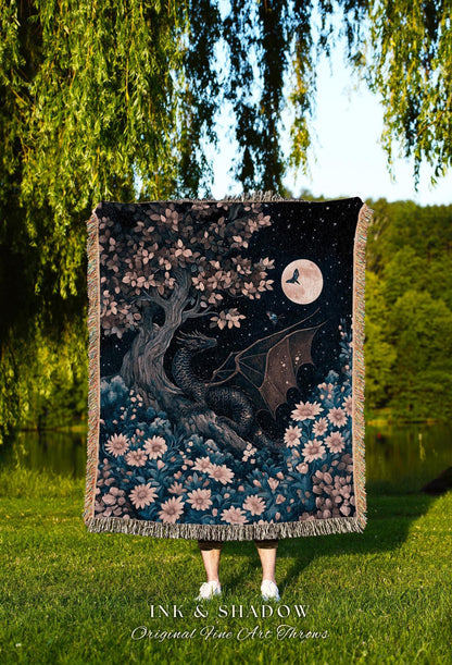 May include: A woven throw blanket featuring a fantasy scene with a black dragon, a full moon, and a tree with pink and white flowers. The blanket has a fringed edge and the text "INK & SHADOW Original Fine Art Throws" at the bottom.