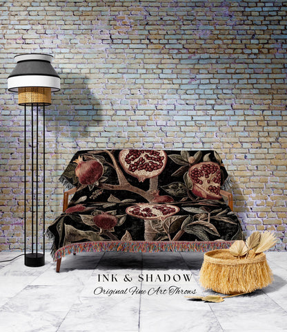 May include: A black throw blanket with a pomegranate design draped over a wooden sofa. The blanket features red and brown pomegranates and foliage. A floor lamp with a black base and a woven shade stands to the left. A woven basket with fringe sits on the floor.