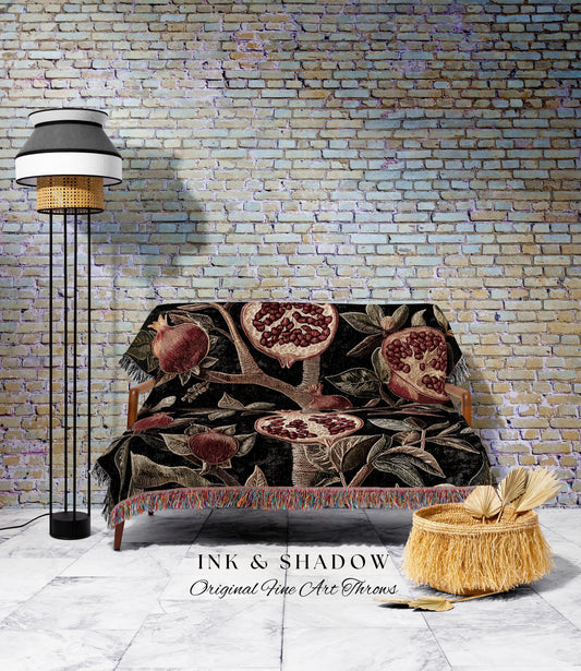 May include: A black throw blanket with a pomegranate design draped over a wooden sofa. The blanket features red and brown pomegranates and foliage. A floor lamp with a black base and a woven shade stands to the left. A woven basket with fringe sits on the floor.