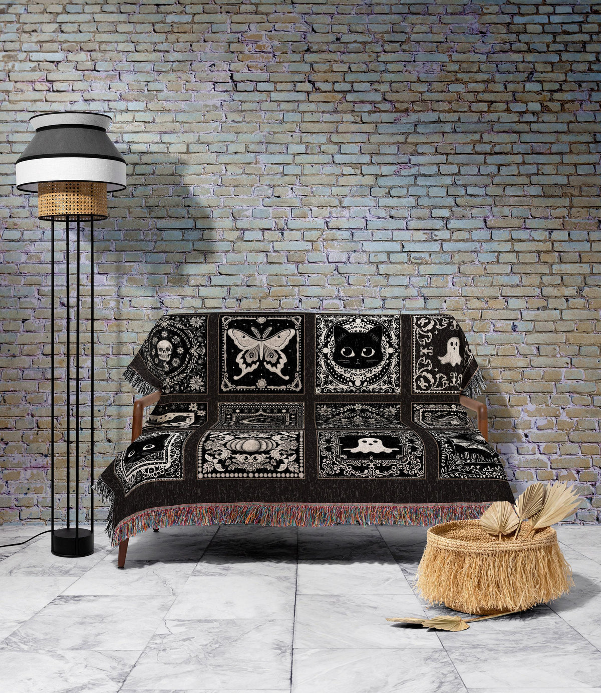 May include: A black and white throw blanket with a repeating pattern of gothic and Halloween-themed squares, including a skull, butterfly, cat, and ghost. The blanket is draped over a wooden sofa. A floor lamp and a woven basket are in the background.