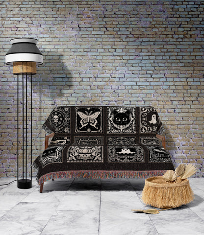 May include: A black and white throw blanket with a repeating pattern of gothic and Halloween-themed squares, including a skull, butterfly, cat, and ghost. The blanket is draped over a wooden sofa. A floor lamp and a woven basket are in the background.