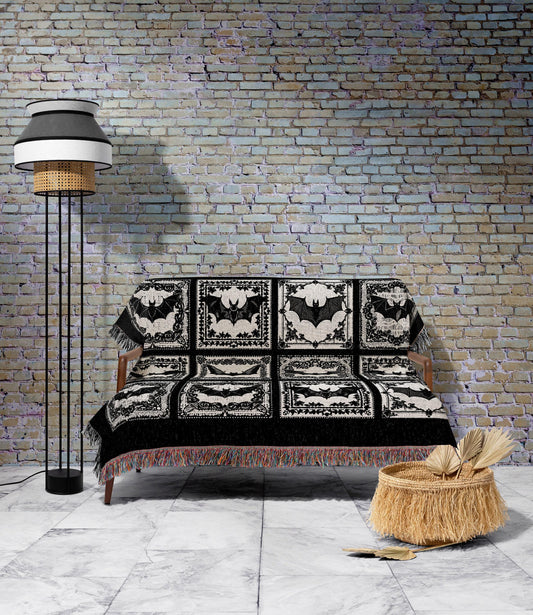 May include: A black and white throw blanket with a bat design draped over a wooden sofa. The blanket features a repeating pattern of bats within decorative frames. A black fringed border and a floor lamp with a black and white shade are also visible.