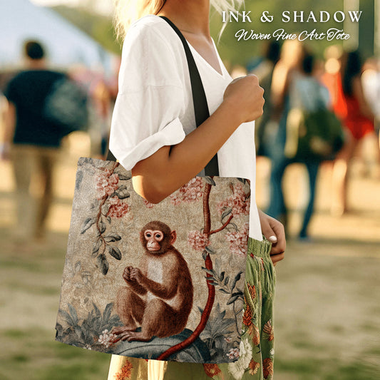 May include: A woven tote bag with a brown monkey sitting on a branch with pink flowers. The bag has the text "INK & SHADOW Woven Fine Art Tote" printed on it.