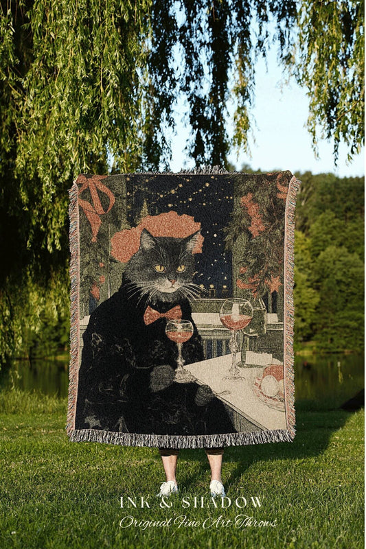 May include: A woven throw blanket featuring a black cat in a tuxedo, holding a wine glass. The cat is depicted in a restaurant setting with two wine glasses and a plate of food. The blanket has a fringed edge and the text "INK & SHADOW Original Fine Art Throws".