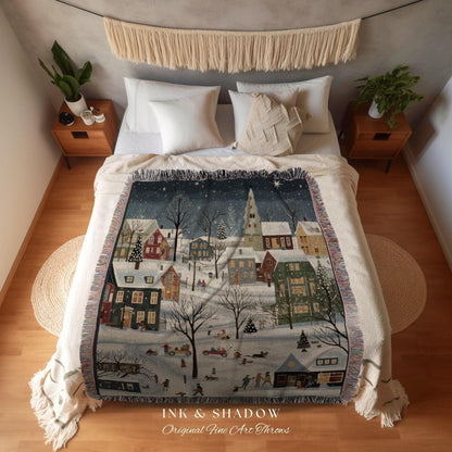 May include: A woven throw blanket featuring a winter village scene with snow-covered houses, trees, and people. The blanket has a cream-colored fringe and the text "INK & SHADOW Original Fine Art Throws" at the bottom.