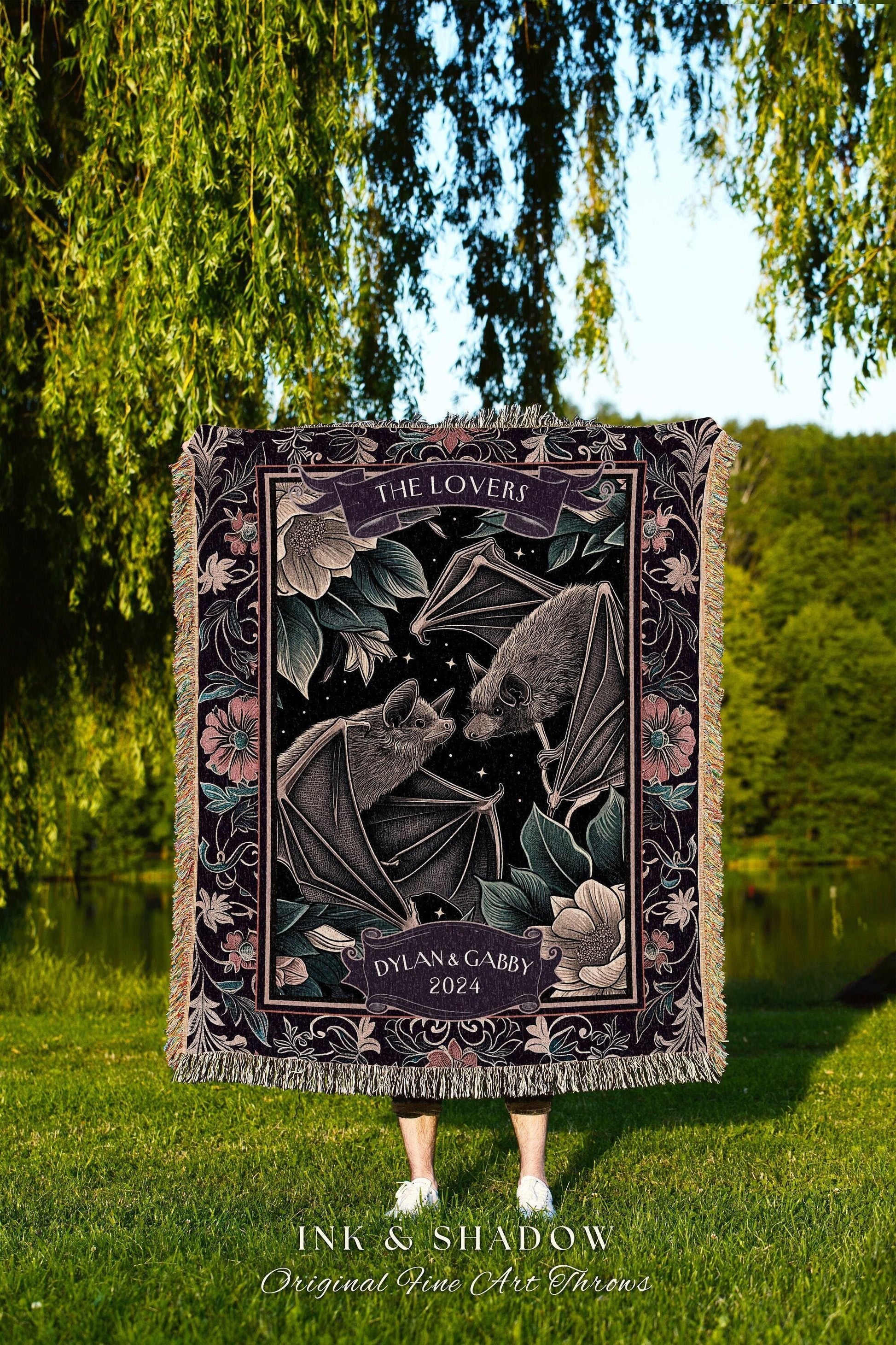 May include: A woven throw blanket with a dark background featuring two bats, flowers, and the text "THE LOVERS" and "DYLAN & GABBY 2024." The blanket has a fringed edge and the words "INK & SHADOW Original Fine Art Throws" are at the bottom.