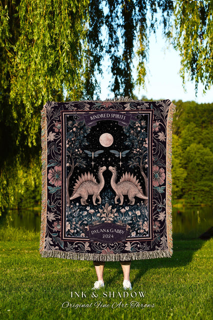 May include: A dark purple throw blanket with a detailed illustration of two dinosaurs facing each other under a full moon. The blanket has a floral border and the words "Kindred Spirits" and "Dylan & Gabby 2024".