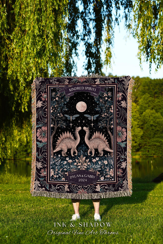 May include: A dark purple throw blanket with a detailed illustration of two dinosaurs facing each other under a full moon. The blanket has a floral border and the words "Kindred Spirits" and "Dylan & Gabby 2024".