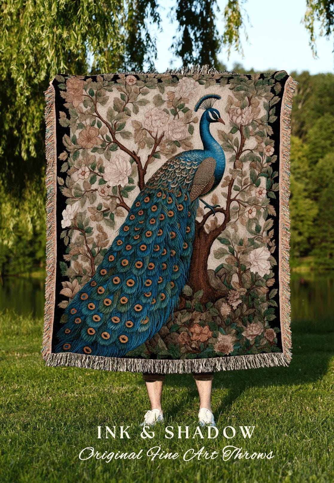 May include: A decorative throw blanket featuring a detailed illustration of a peacock perched on a tree branch, surrounded by floral elements. The peacock has vibrant blue and green feathers. The blanket has a black border and fringe.