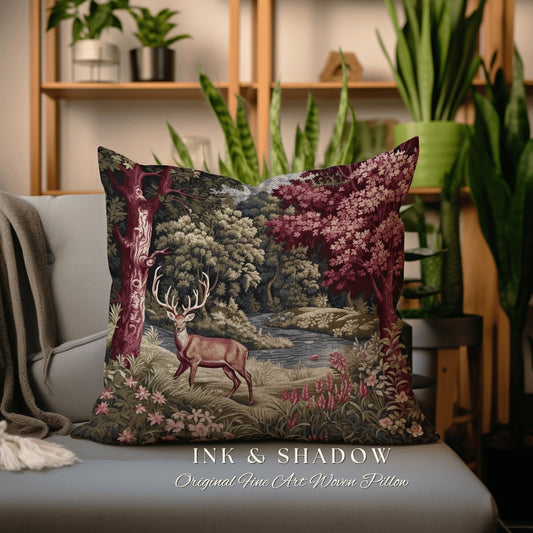 May include: A decorative pillow featuring a woven tapestry design of a deer standing in a forest with a river. The pillow has a dark background with shades of green, brown, and red. The text "INK & SHADOW Original Fine Art Woven Pillow" is printed on the bottom of the pillow.