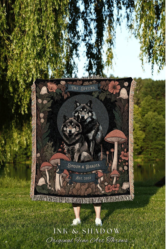 May include: A woven throw blanket featuring a detailed illustration of two wolves, surrounded by mushrooms and floral elements. The blanket has a dark background with the text "The Lovers", "London & Dakota", and "Est. 2023". The blanket is fringed.