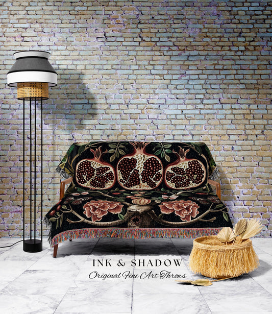 May include: A black throw blanket with a pomegranate design is draped over a wooden sofa. The blanket features red, brown, and green accents. A floor lamp with a black base and a woven shade stands to the left. A woven basket with fringe sits on the floor.
