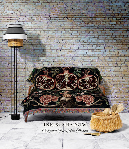 May include: A black throw blanket with a pomegranate design is draped over a wooden sofa. The blanket features red, brown, and green accents. A floor lamp with a black base and a woven shade stands to the left. A woven basket with fringe sits on the floor.