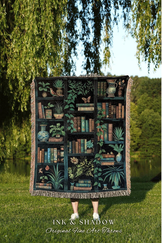 May include: A black throw blanket featuring a detailed illustration of a bookshelf filled with books, plants, and decorative vases. The design includes a variety of green plants and a butterfly. The blanket has a fringed edge and the text "INK & SHADOW Original Fine Art Throws".