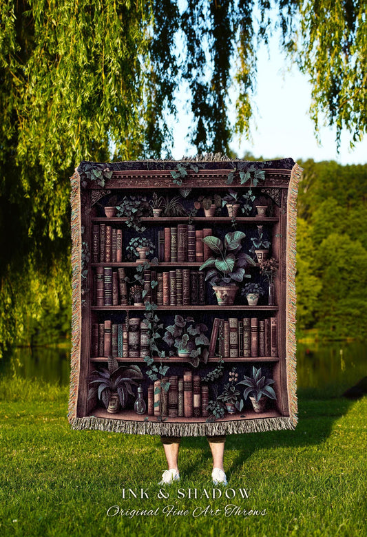 May include: A throw blanket featuring a detailed illustration of a bookshelf filled with books and potted plants. The blanket has a fringed edge and the text "INK & SHADOW Original Fine Art Throws" at the bottom.