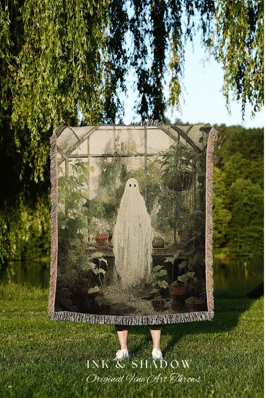 May include: A woven throw blanket featuring a ghost in a greenhouse scene. The artwork includes potted plants and a glass structure. The blanket has a fringed edge and the text "INK & SHADOW Original Fine Art Throws".