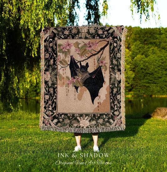 May include: A tapestry throw blanket featuring a bat hanging from a flowering branch. The bat is black, and the flowers are pink. The border is dark green with floral patterns. The text "INK & SHADOW Original Fine Art Throws" is at the bottom.