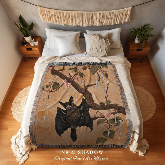 May include: A beige throw blanket featuring a bat with outstretched wings, perched on a flowering tree branch. The design includes pink blossoms and green leaves against a tan background. The blanket has a fringed edge and the text "INK & SHADOW Original Fine Art Throws".