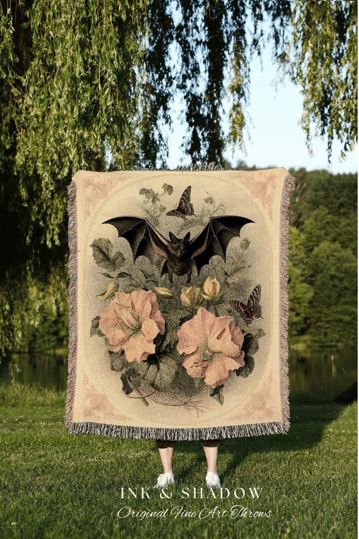 May include: A beige throw blanket featuring a bat with outstretched wings, butterflies, and large pink flowers. The blanket has a fringed edge and the text "INK & SHADOW Original Fine Art Throws" at the bottom.