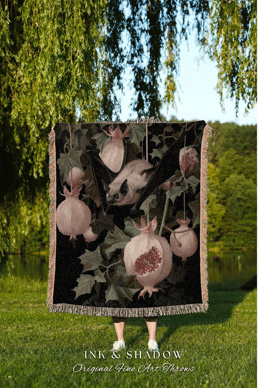 May include: A decorative throw blanket featuring a bat, pomegranates, and foliage in shades of black, green, and pink. The blanket has a fringed edge and the text "INK & SHADOW Original Fine Art Throws" at the bottom.