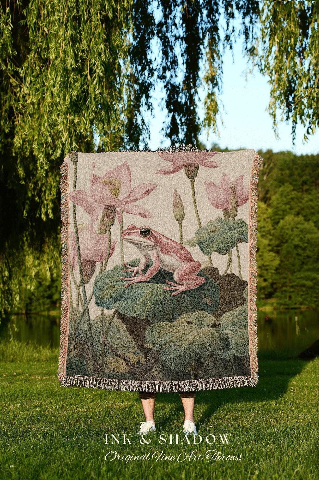 May include: A woven throw blanket featuring a pink frog on a lily pad, surrounded by pink lotus flowers and green leaves. The blanket has a fringed edge and the text "INK & SHADOW Original Fine Art Throws" at the bottom.