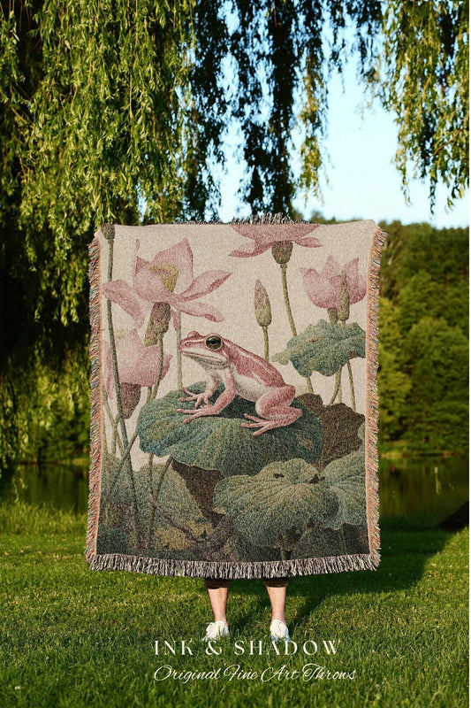 May include: A woven throw blanket featuring a pink frog on a lily pad, surrounded by pink lotus flowers and green leaves. The blanket has a fringed edge and the text "INK & SHADOW Original Fine Art Throws" at the bottom.