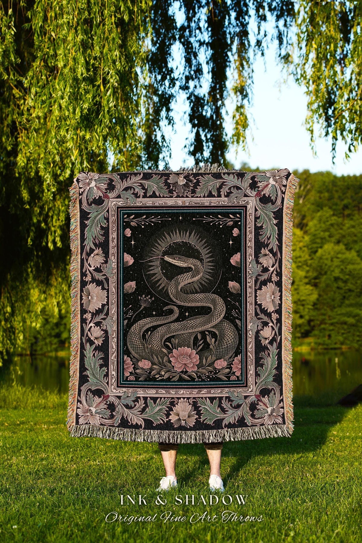 May include: A black throw blanket with a detailed illustration of a snake, flowers, and celestial elements. The blanket has a fringed edge and the text "INK & SHADOW Original Fine Art Throws". The design is in shades of gray, pink, and black.