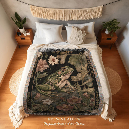 May include: A woven throw blanket featuring a detailed illustration of a green frog amidst flowers and foliage on a black background. The blanket has a fringed edge and the text "INK & SHADOW Original Fine Art Throws".