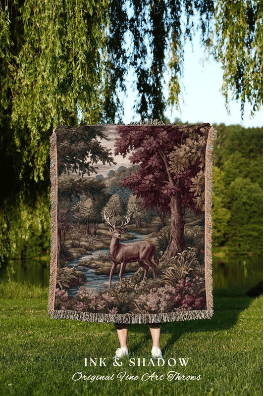 May include: A tapestry throw blanket featuring a detailed forest scene with a deer, river, and trees in shades of brown, green, and burgundy. The blanket has a fringed edge and the text "INK & SHADOW Original Fine Art Throws".