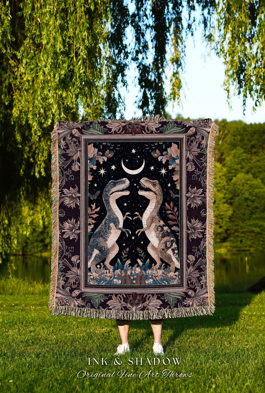 May include: A tapestry throw blanket featuring two Tyrannosaurus Rex dinosaurs facing each other under a crescent moon and stars. The blanket has a dark background with floral and leaf accents, and a fringed edge. The text "INK & SHADOW Original Fine Art Throws" is at the bottom.