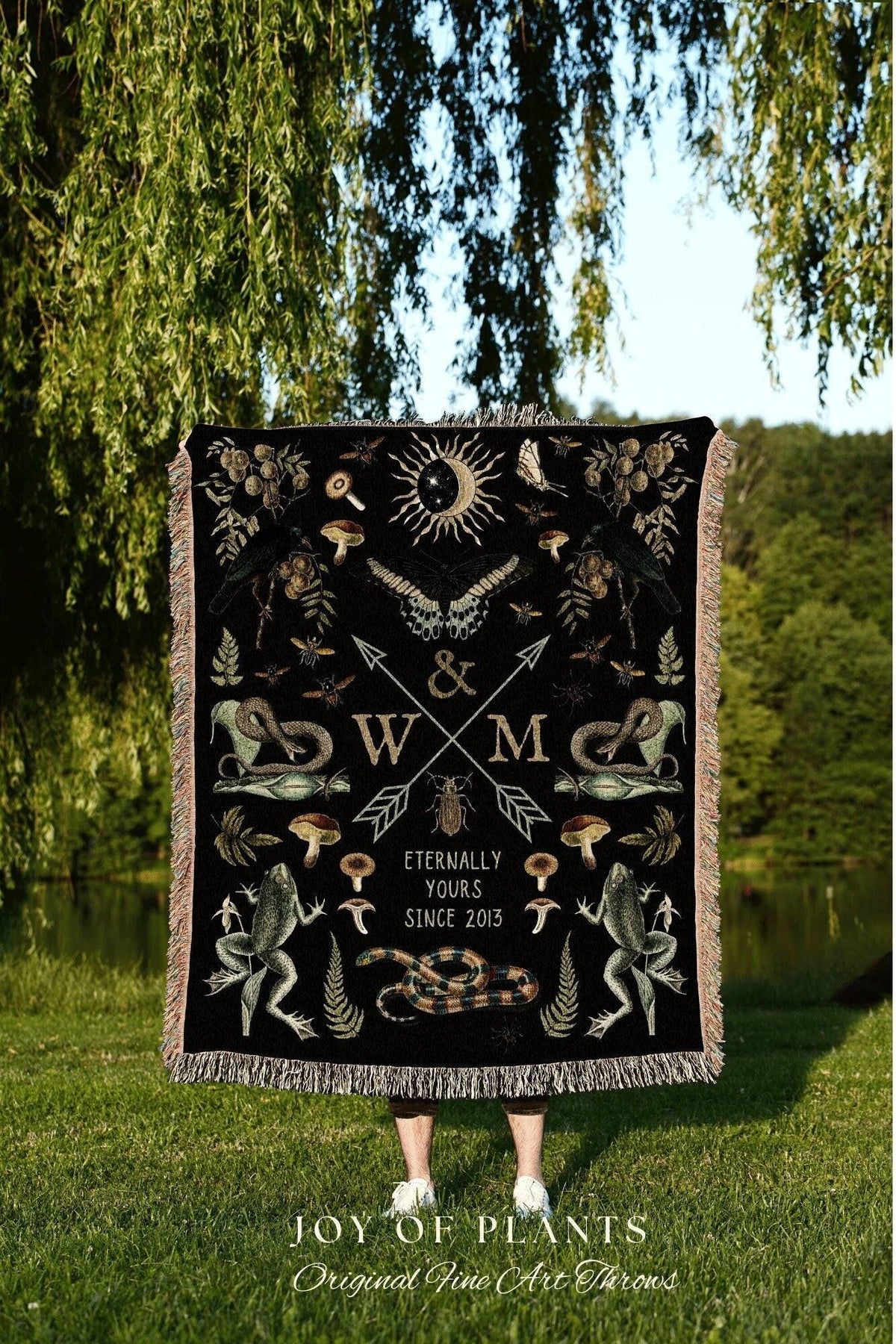 May include: A black throw blanket with a detailed design featuring a sun, moon, butterflies, snakes, frogs, and mushrooms. The blanket has a fringe border and the text "Eternally Yours Since 2013". The blanket is held up in front of a person.