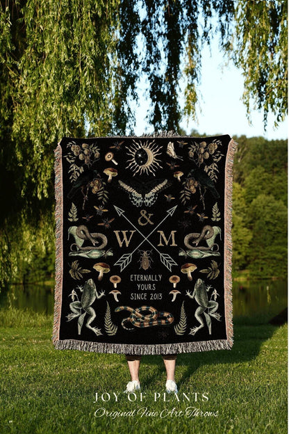 May include: A black throw blanket with a detailed design featuring a sun, moon, butterflies, snakes, frogs, and mushrooms. The blanket has a fringe border and the text "Eternally Yours Since 2013". The blanket is held up in front of a person.