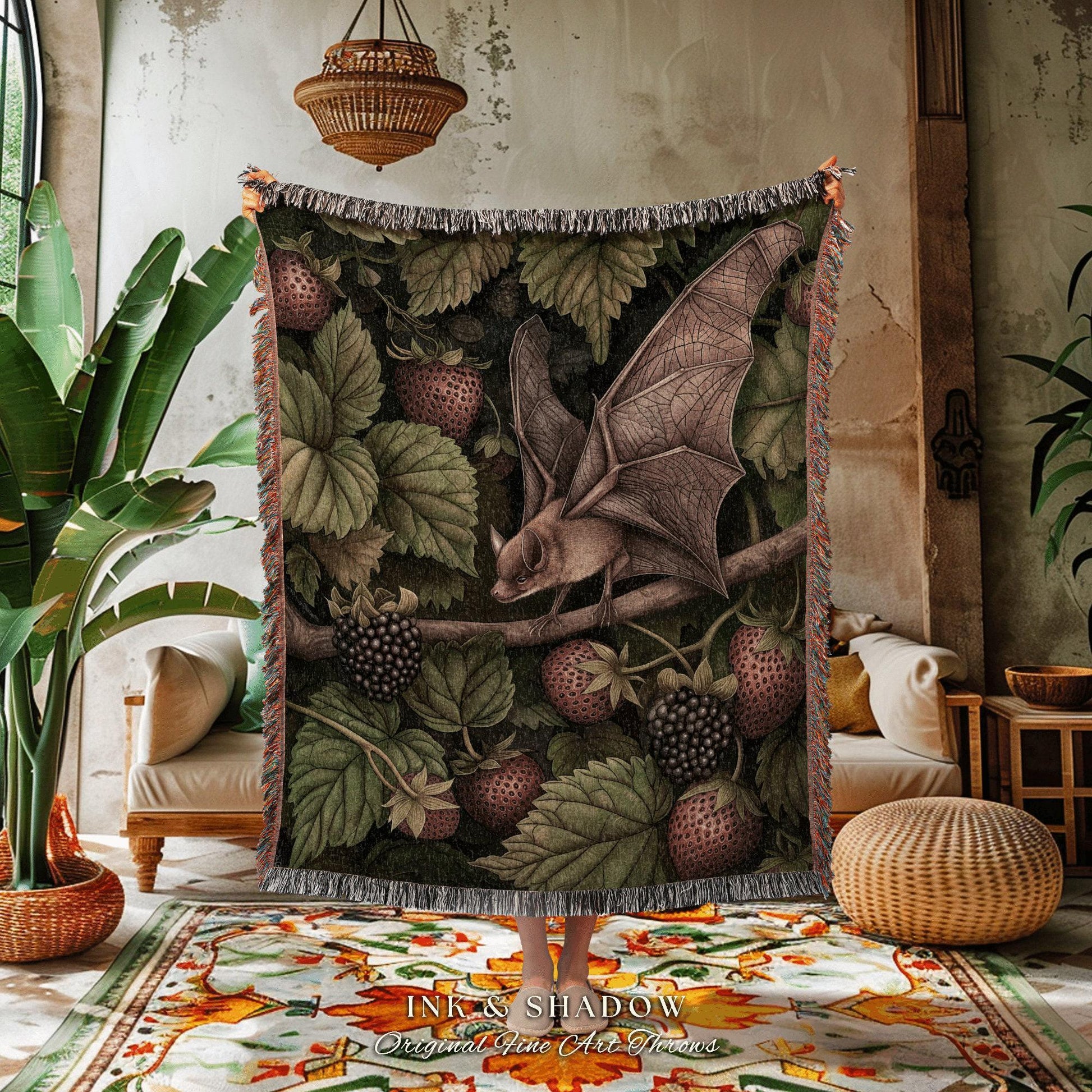 May include: A woven throw blanket featuring a detailed illustration of a bat perched on a branch amidst strawberries and blackberries. The blanket has a dark green background with a fringed edge. The text "INK & SHADOW Original Fine Art Throws" is at the bottom.