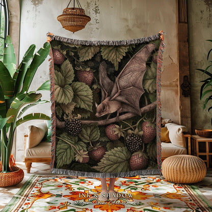 May include: A woven throw blanket featuring a detailed illustration of a bat perched on a branch amidst strawberries and blackberries. The blanket has a dark green background with a fringed edge. The text "INK & SHADOW Original Fine Art Throws" is at the bottom.