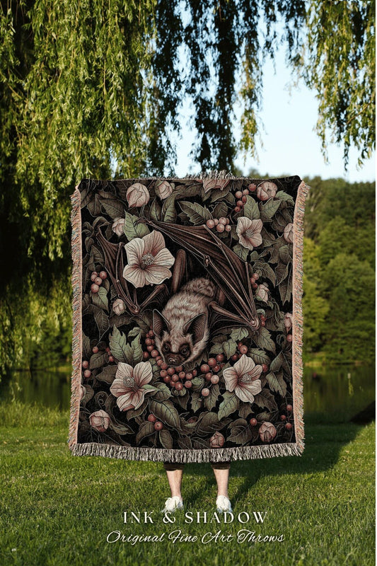May include: A decorative throw blanket featuring a bat surrounded by flowers and berries. The design is in shades of brown, green, and pink, with a fringed edge. The blanket has the text "INK & SHADOW Original Fine Art Throws" at the bottom.