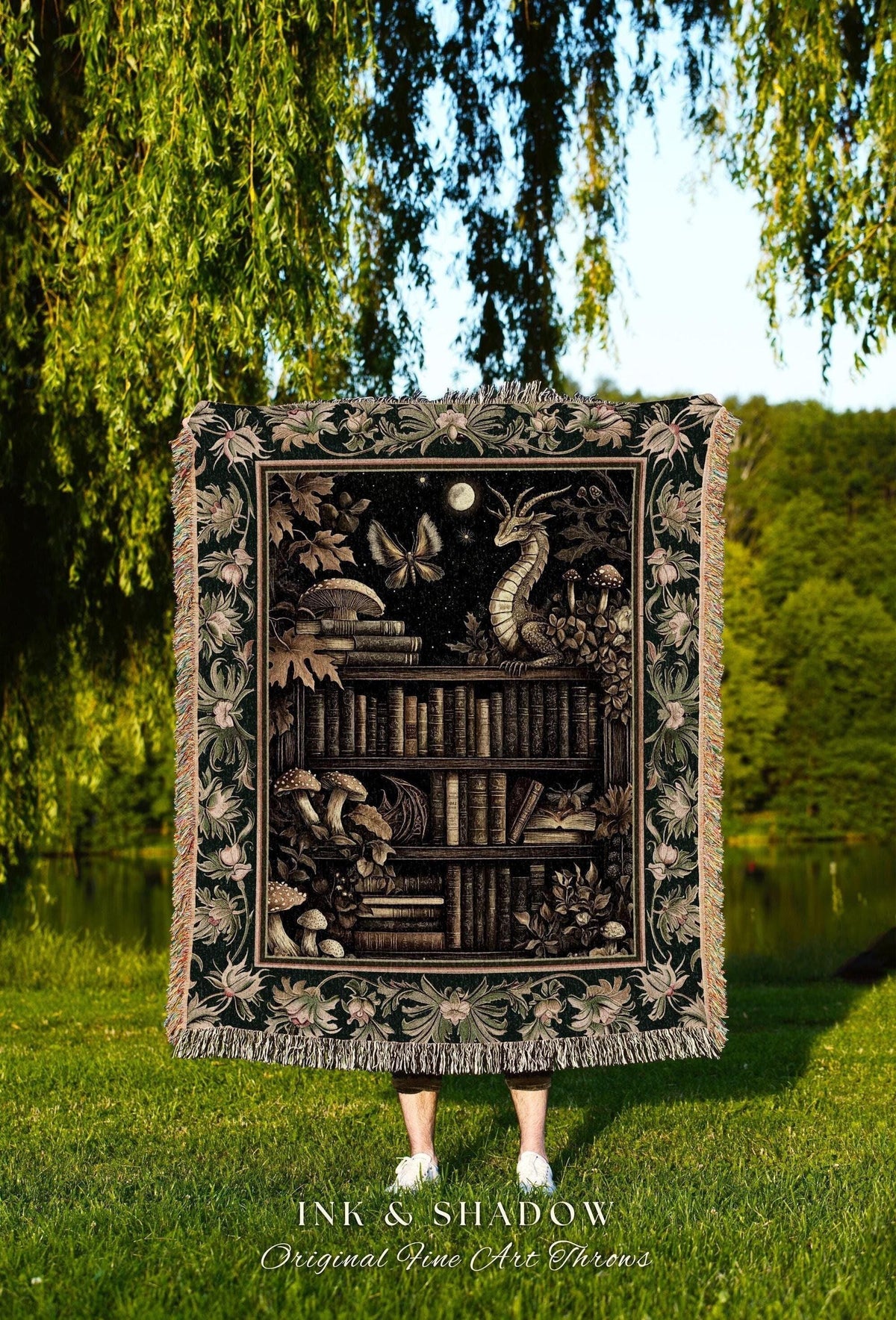 May include: A dark green throw blanket with a fantasy bookshelf design. The design includes a dragon, butterflies, mushrooms, and a moon. The blanket has a fringed edge and the text "INK & SHADOW Original Fine Art Throws".