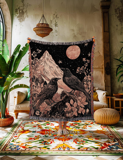 May include: Black woven throw blanket featuring a mountain scene with two crows perched on a branch under a starry night sky and a large pink moon. The blanket has a fringed edge and the text "INK & SHADOW Original Fine Art Throws".