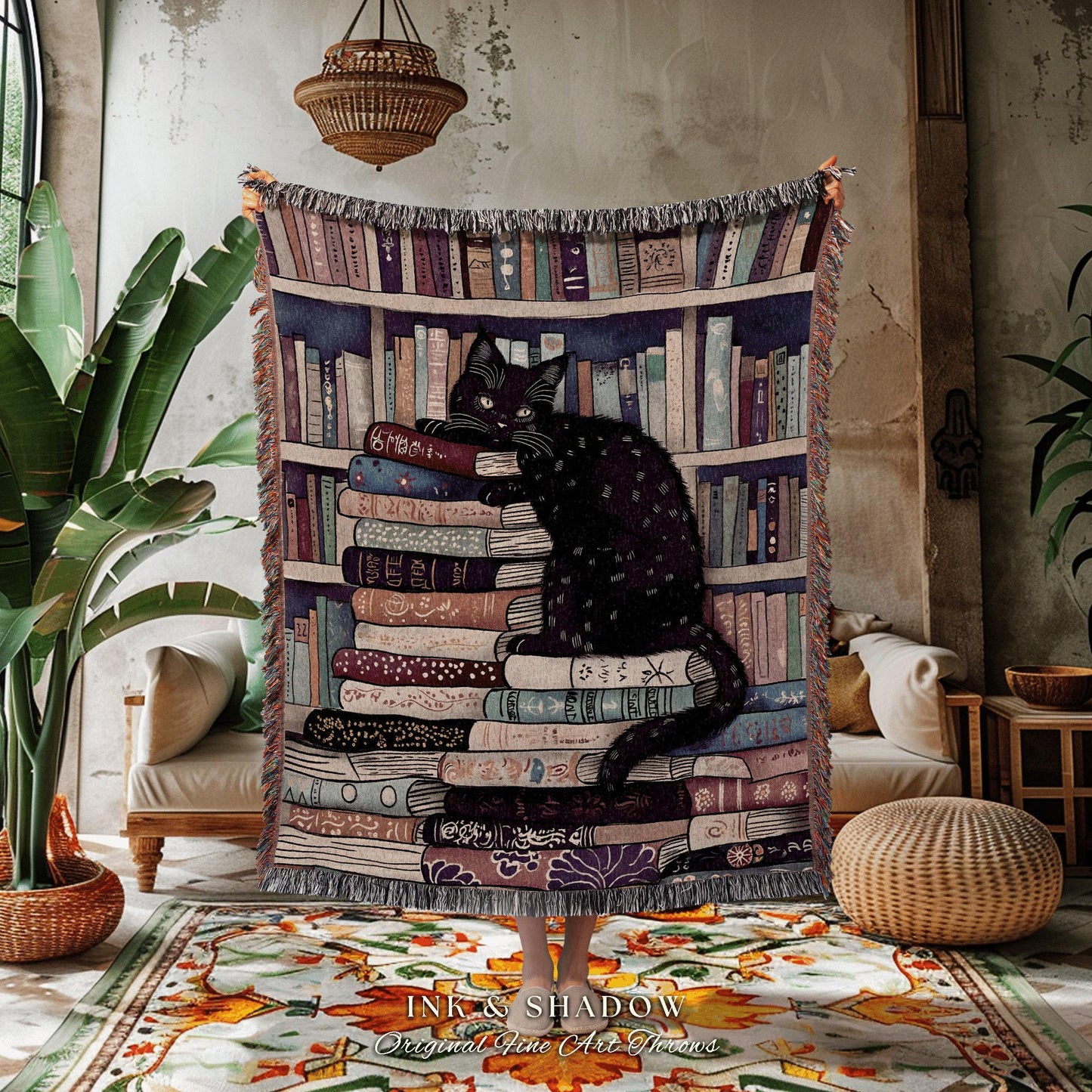 May include: A decorative throw blanket featuring a black cat perched atop a stack of books in front of a bookshelf backdrop. The blanket has a fringed edge and the text "INK & SHADOW Original Fine Art Throws" at the bottom.