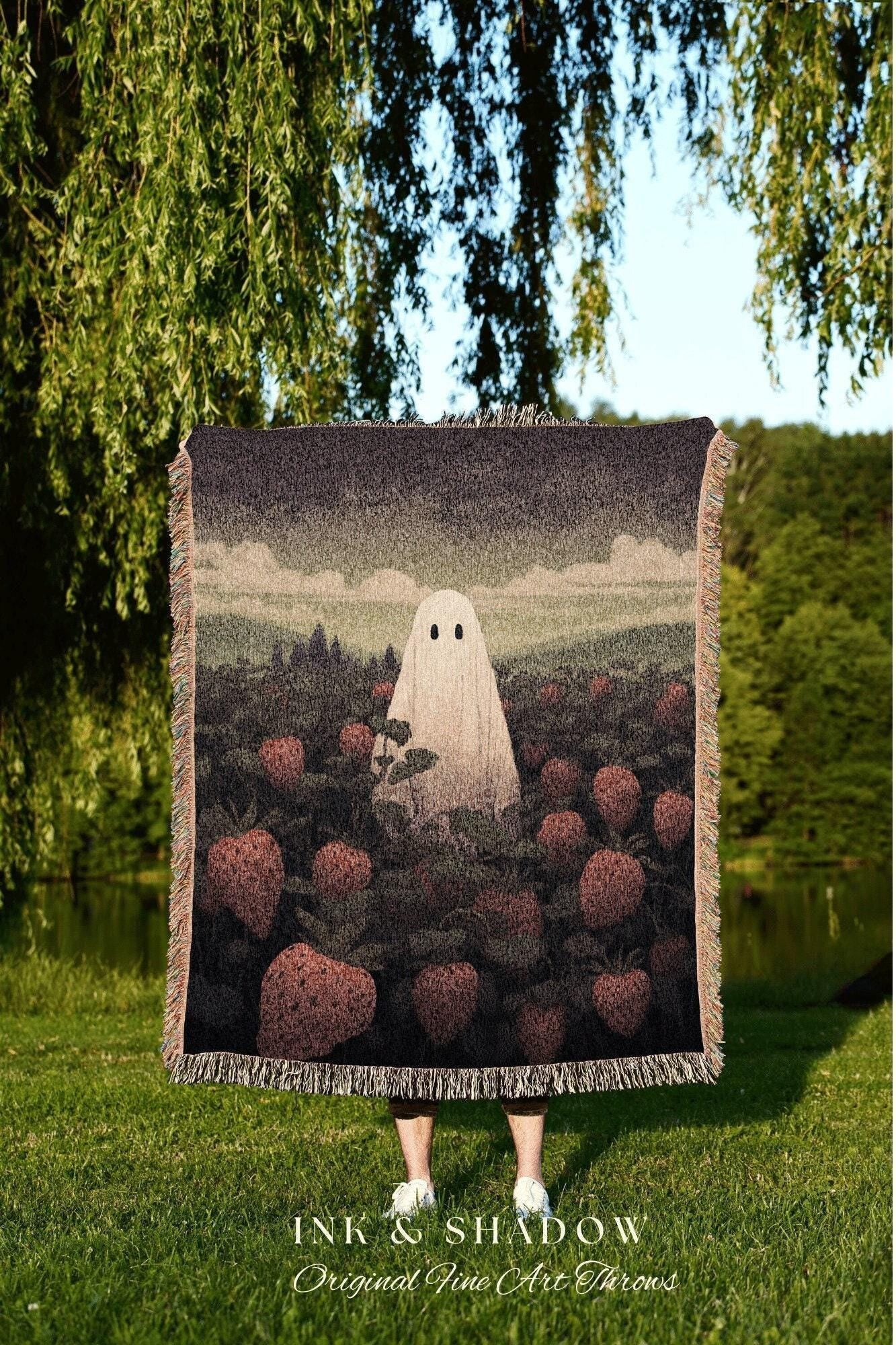 May include: A woven throw blanket featuring a whimsical design of a ghost in a field of strawberries. The blanket has a dark background with a fringe trim and the text "INK & SHADOW Original Fine Art Throws".