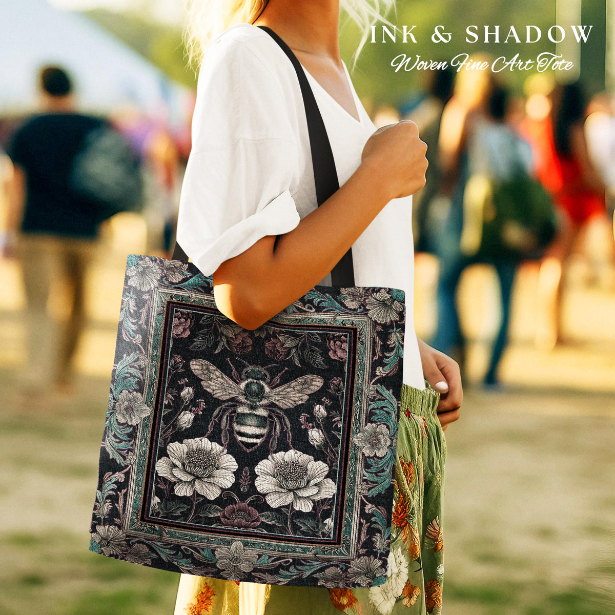 May include: A black tote bag with a floral and bee design. The bag has a black strap and is made of woven fabric. The text "INK & SHADOW Woven Fine Art Tote" is printed on the bag.