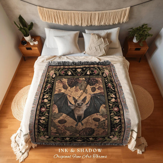 May include: A decorative throw blanket featuring a bat with outstretched wings, surrounded by botanical elements. The blanket has a black border with floral patterns and the text "INK & SHADOW Original Fine Art Throws". The color palette includes black, beige, and shades of green.