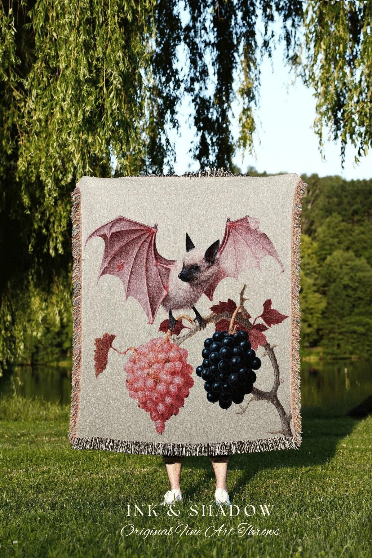 May include: A beige throw blanket with a bat and grape illustration. The bat has pink wings and is perched on a branch with pink and black grapes. The blanket has a fringed edge and the text "INK & SHADOW Original Fine Art Throws".