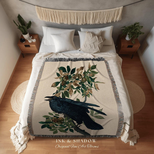 May include: A cream-colored throw blanket featuring a detailed illustration of a black raven perched on a branch with green leaves. The blanket has a fringed edge and the text "INK & SHADOW Original Fine Art Throws" is printed at the bottom.