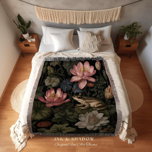 May include: A woven throw blanket featuring a botanical design with pink lotus flowers, green lily pads, and a frog on a black background. The blanket has a cream-colored fringe and the text "INK & SHADOW Original Fine Art Throws".