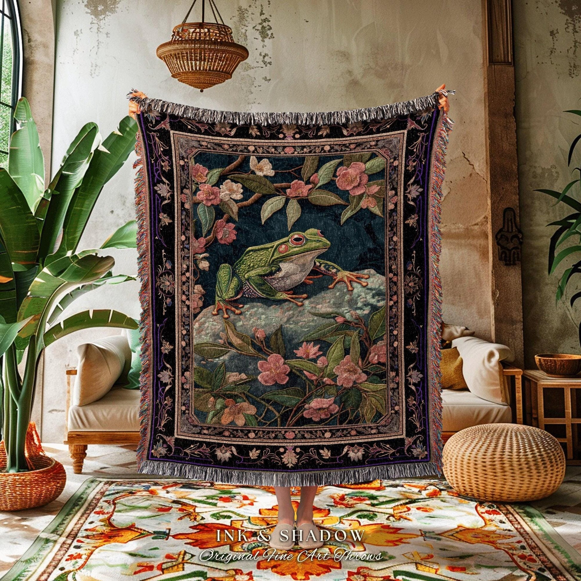 May include: A tapestry throw blanket featuring a green frog on a rock surrounded by pink flowers and foliage. The blanket has a dark blue background and a decorative border with fringe. The text "INK & SHADOW Original Fine Art Home" is at the bottom.