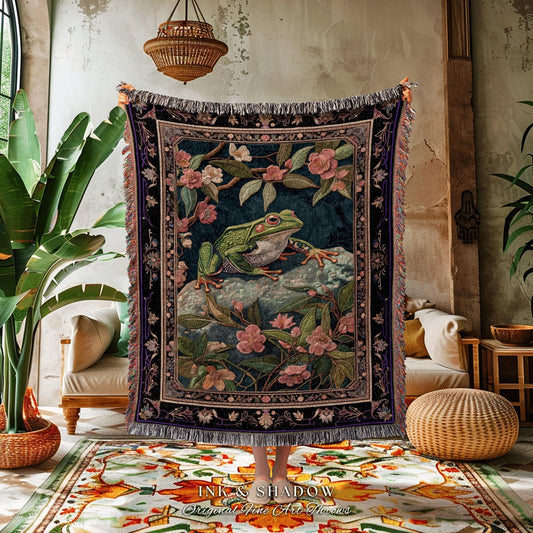 May include: A tapestry throw blanket featuring a green frog on a rock surrounded by pink flowers and foliage. The blanket has a dark blue background and a decorative border with fringe. The text "INK & SHADOW Original Fine Art Home" is at the bottom.